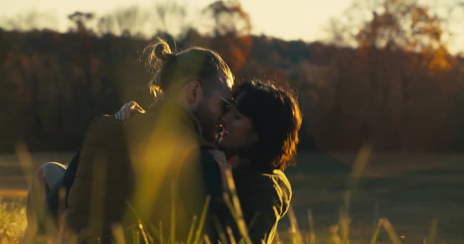 A couple is kissing while sitting in a grassy field during sunset
