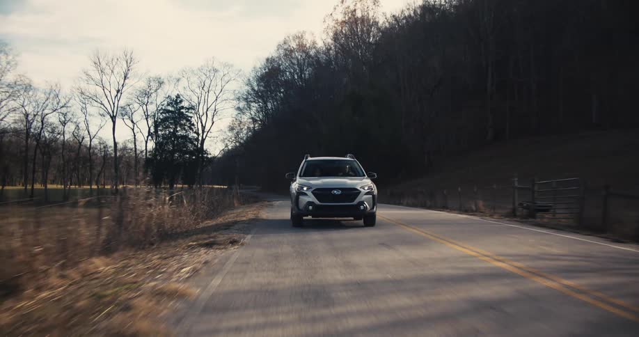 A gray suv is driving towards the camera on a twolane country road with trees and grassy fields on either side