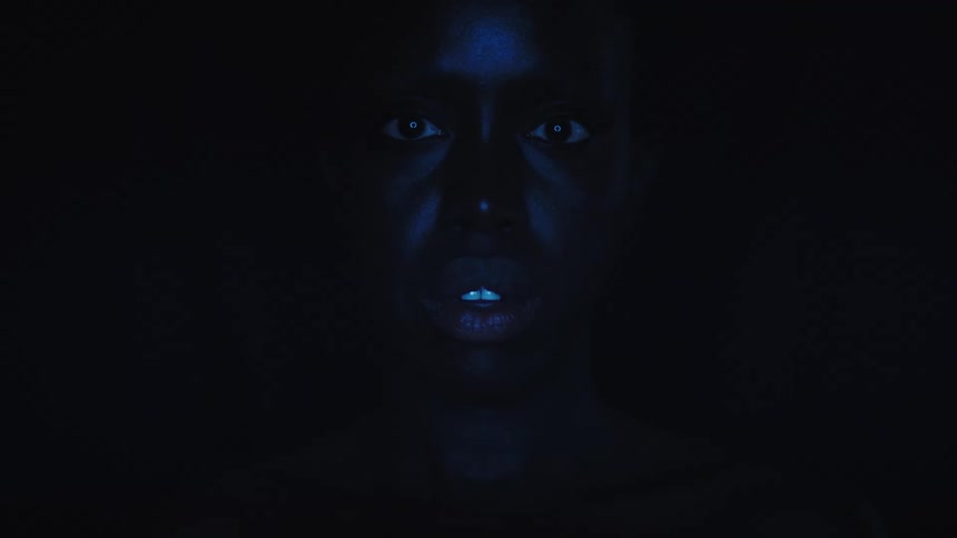 A woman face is dimly lit by a blue light revealing his eyes and mouth as he stares directly at the camera in a dark room