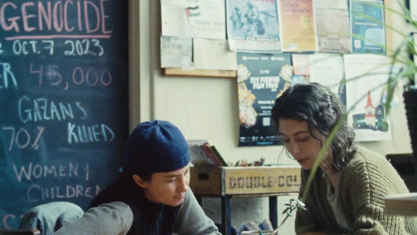 A female teacher instructs a boy student both seated at the front of the class with a blackboard behind them at midday