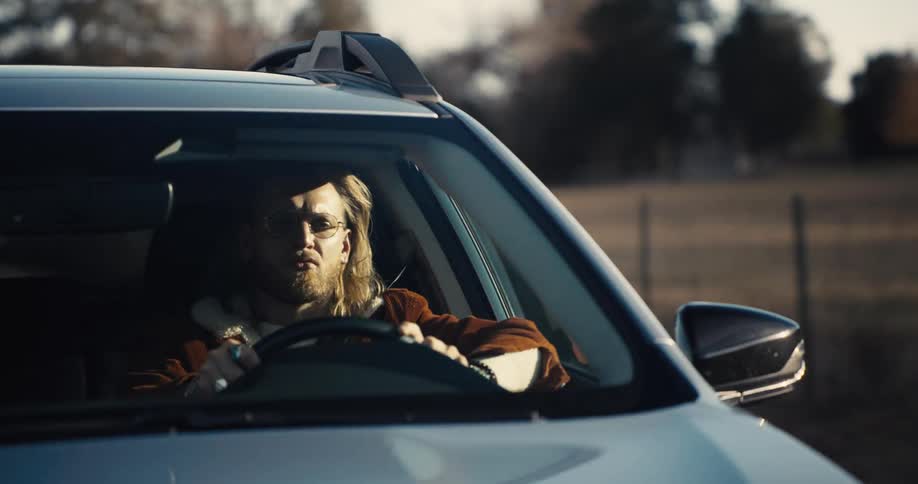 A man with long light colored hair and a beard sits in the drivers seat of a car wearing glasses and a furry collared jacket
