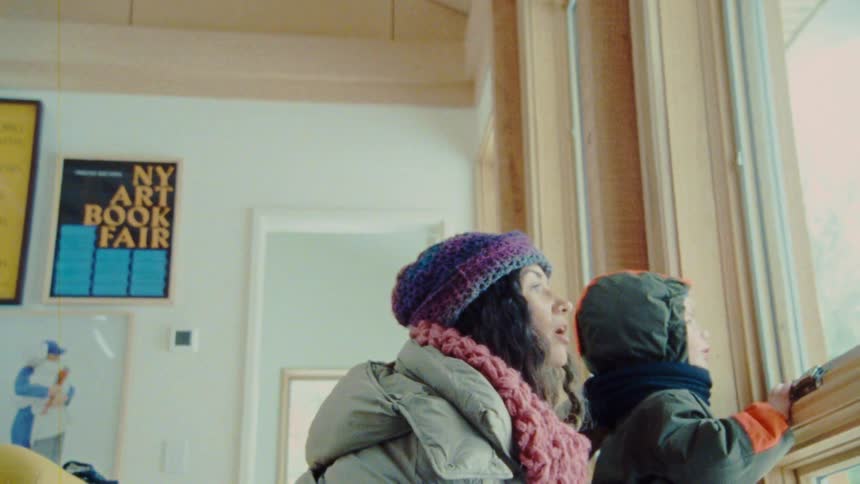 A woman and a child stand looking out a large window in a room with a high wood paneled ceiling with the woman wearing a purple knit hat and pink scarf and the child wearing a green hat and orange scarf