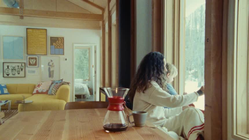 A woman and a child sit on a window sill with their backs to the camera looking out at a snowy outdoor scene as a glass coffee carafe and mug sit on a wooden table in the foreground
