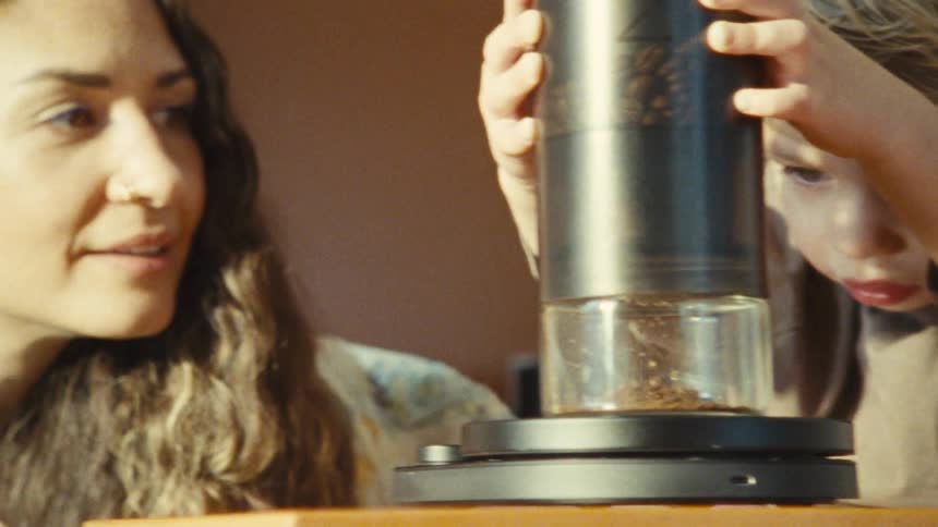 A young child with light colored hair and a brown shirt stands with a woman as the child holds a coffee grinder and watches the ground beans fall into the glass canister