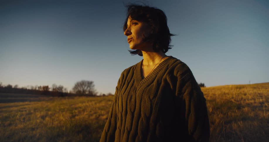A woman with dark hair and a chunky brown sweater stands in a dry grassy field during the golden hour looking off to her left as the sun sets behind her