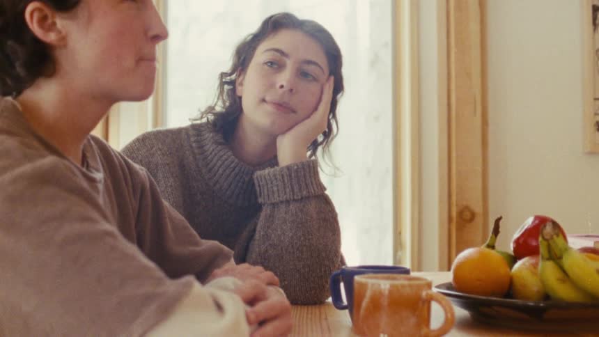 A young woman is resting her chin on her hand while listening intently to another woman at a wooden table in a kitchen setting