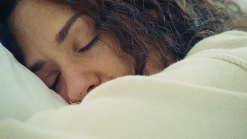 The video shows a closeup of a person with curly dark hair sleeping peacefully on a pillow their eyes are closed and they have a gentle serene smile on their face