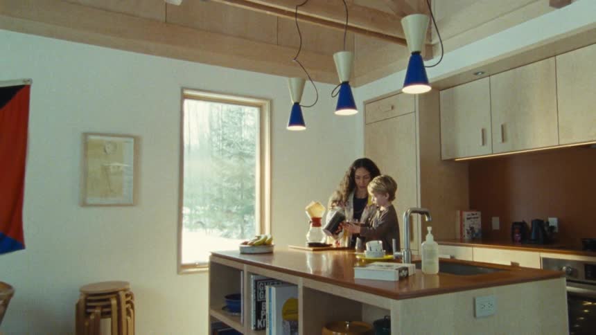 A woman with a boy in the kitchen pouring something into his cup while he drinks