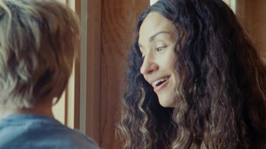A close view shows a woman with long dark curly hair smiling and talking to a child with light colored hair then she extends her hands and wiggles her fingers playing a game