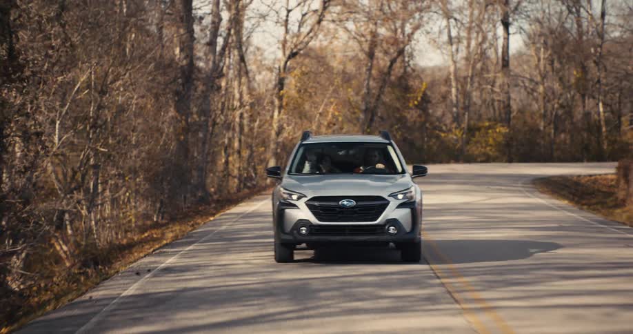 A silver subaru outback wagon drives on a winding road through a forest with dry bare trees