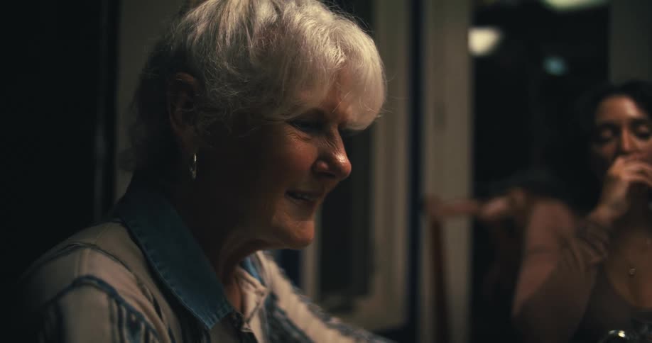 A woman with white hair and a white and blue striped shirt smiles and talks with a young woman eating in the background