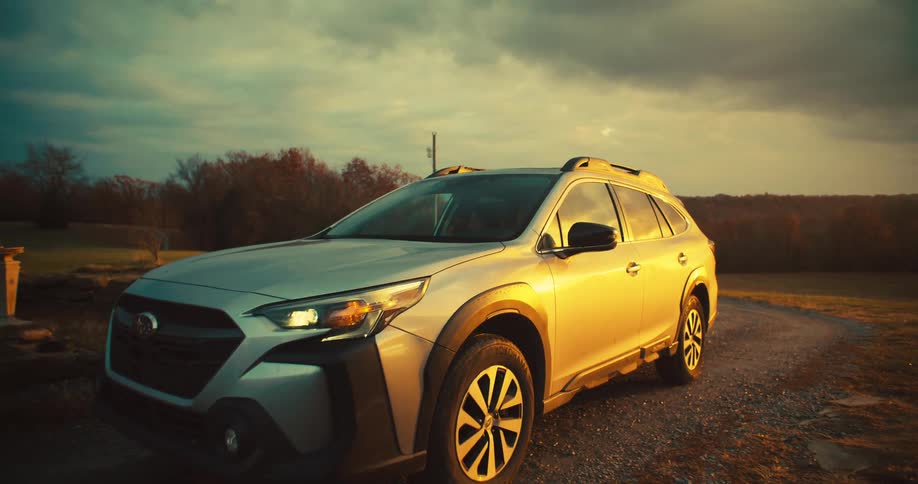 A car with headlights on drives up a gravel path and a person exits the suv