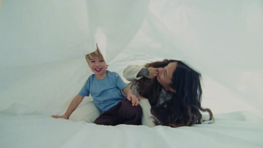 A mother plays with her son on a bed holding a blanket over their heads as the child moves and jumps with excitement