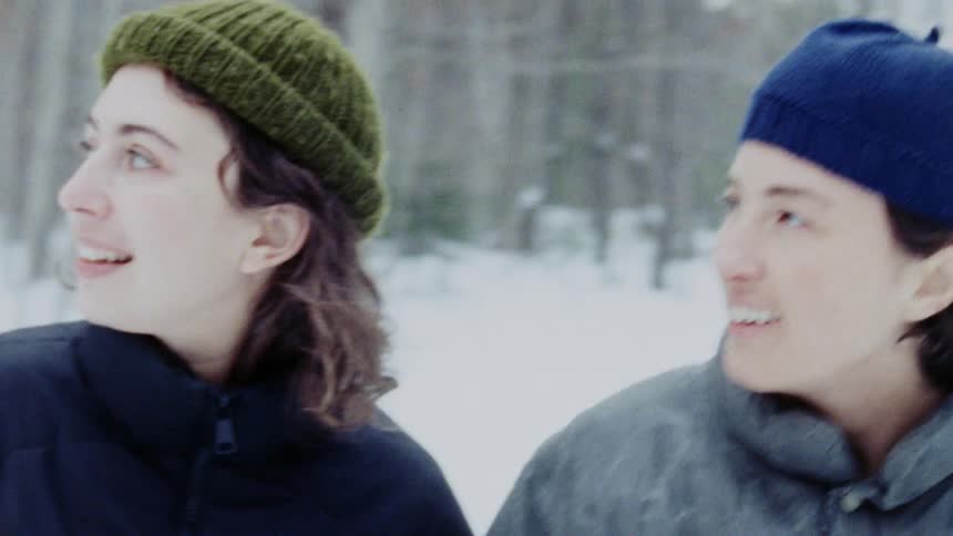 A young couple holds hands as they walk through the snow in the woods wearing winter clothing at midday