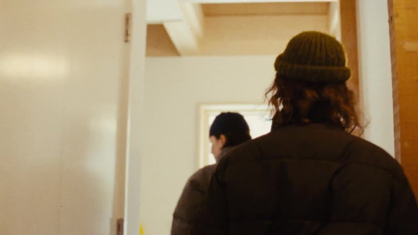 Two people a man with long hair and a knit hat and a woman with a dark hat and jacket walk down a partially renovated hallway with exposed wooden studs on the right