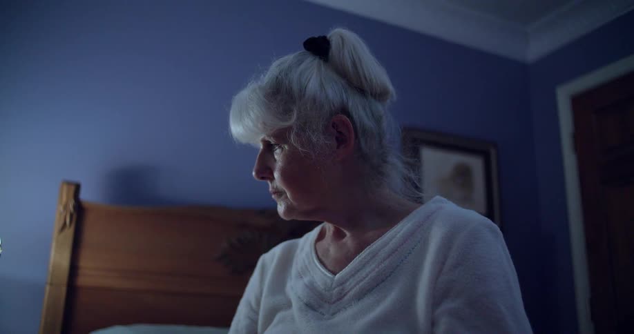 The video shows an elderly woman with white hair sitting on a bed in a dimly lit room she is looking down and appears to be deep in thought