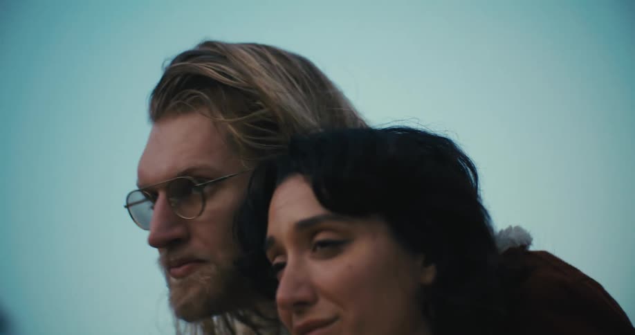 A man and a woman with a sad look on her face stand beside each other as they look into the distance then he kisses her forehead as she smiles and turns to her side revealing a smiling elderly woman
