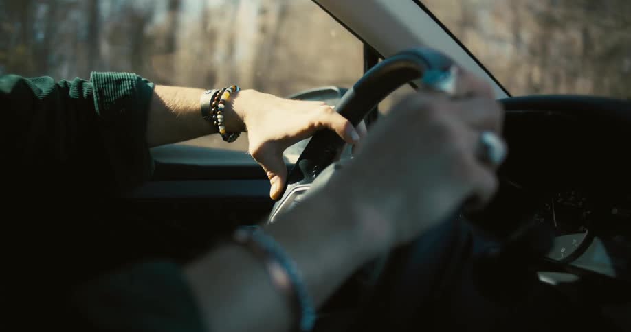 Close up of a vehicle steering wheel as a young man drives on a street during daytime