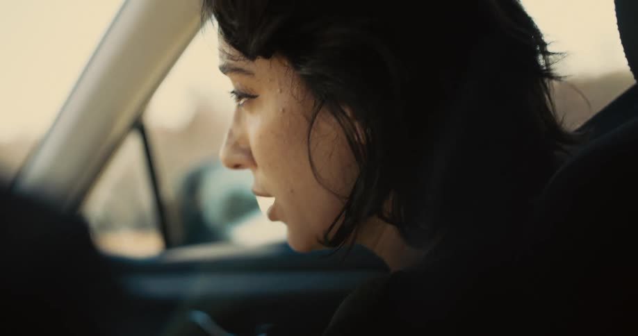A young woman sits in the passenger seat of a moving car as the driver uses his left arm to rub the back of her head at midday