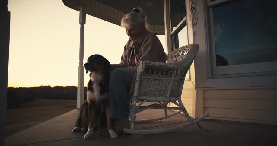 Elderly woman sitting in a rocking chair pets a dog at sunset
