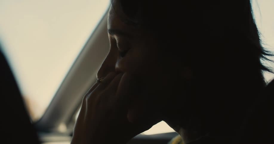 A sad woman rides on a cars passenger seat with her hand over the lower half of her face then she removes it