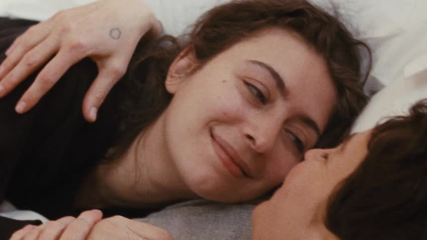 A close view of a woman with dark hair and a light colored ring on her left hand as she lies down next to another person with her eyes closed at first then opens them and smiles as she leans in to kiss the other person