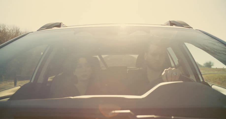 A young man and woman sit in a car as the man drives along a lonely road lined with leafless trees in fall both engaged in conversation at midday