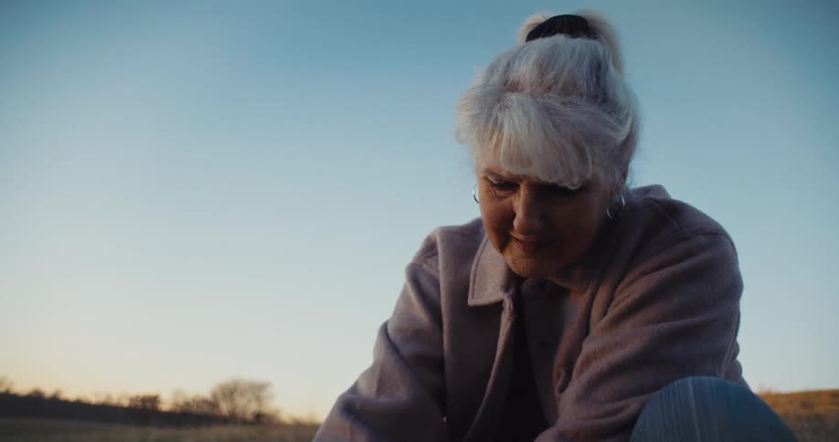 A lowangle shot shows an older woman with white hair pulled back in a ponytail crouching in a field she looks down and then back up with a slight smile as the setting sun casts a warm light on her face the background is a wide clear sky and a field