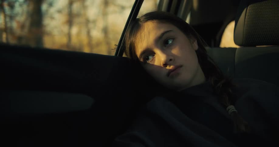 A close view of a woman with her hair in a braid shows her resting her head against the car window and looking out with a neutral expression with the last rays of sun from a golden hour illuminating her face