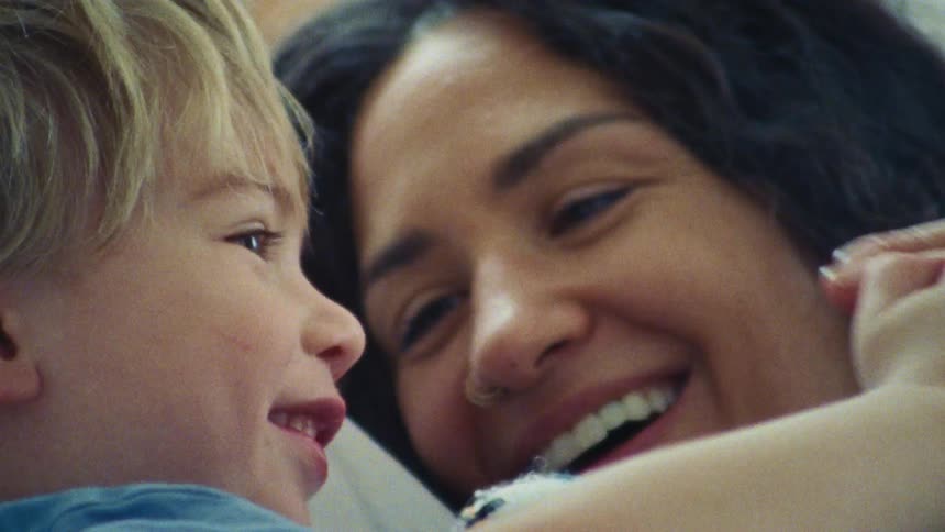 A woman with long curly hair as she touches the hands of a young boy and smiles at him with a joyous expression on both of their faces