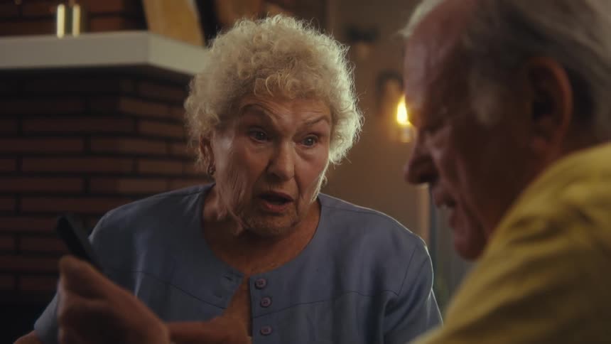 Elderly woman reacts with surprise while looking at something on the phone beside a smiling man