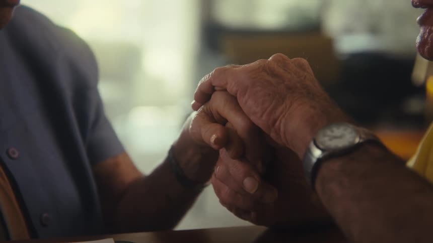 A close view of a man and a woman shows their hands clasped together on a table