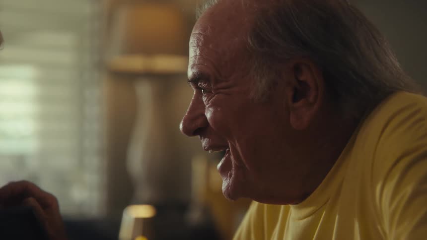 An elderly couple sits in the living room holding hands and laughing during a discussion