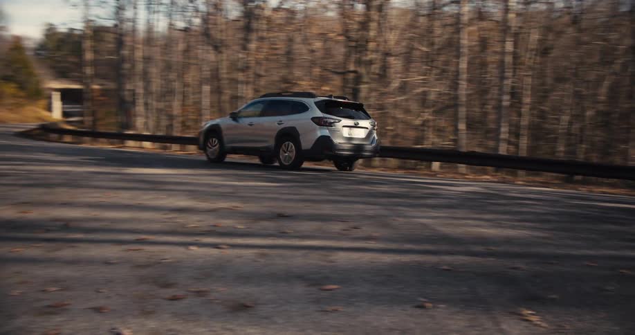 A grey suv drives down a winding road in the woods with fallen leaves on the ground