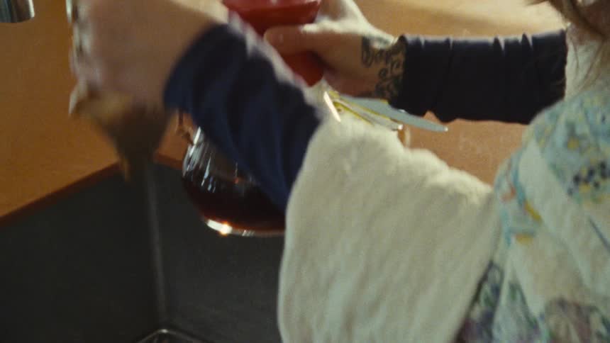 The video shows a closeup of a persons hands holding a glass coffee decanter and pouring the dark liquid into a white mug on a yellow saucer the background is a brightly lit kitchen with a window and a hand reaching for a piece of fruit
