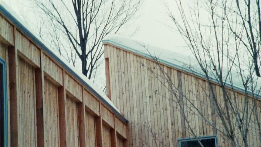 A building with light colored wood siding and a snow covered roofline is seen from a low angle with bare trees and a gray sky in the background