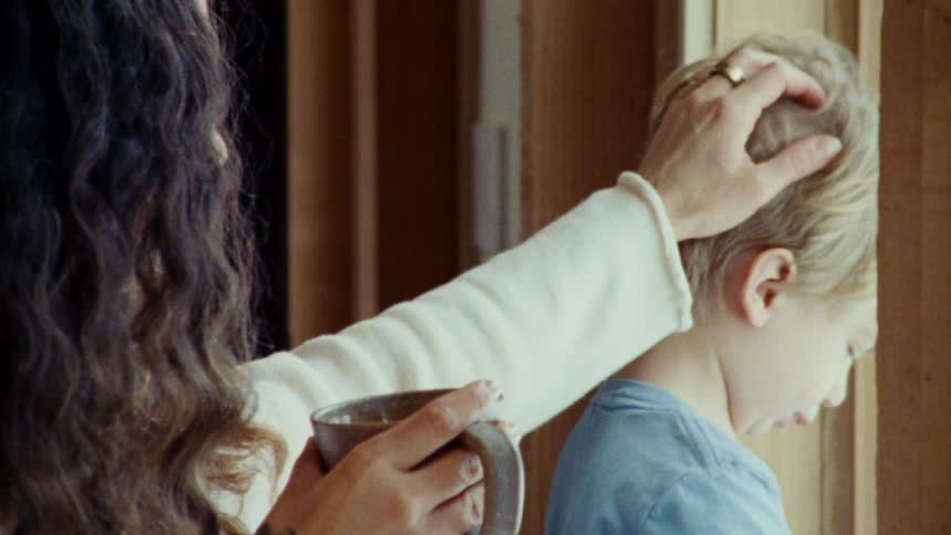 A woman with long dark hair is seen from the side holding a mug in one hand and stroking a childs hair with the other as they both look out a large window