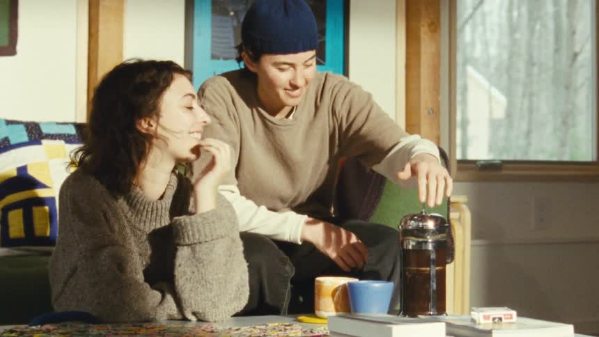 A woman with a beanie approaching a woman from behind who is intently focused on a puzzle on a coffee table and handing her a cup of coffee as she looks up at her and her serious expression changes to a bright smile