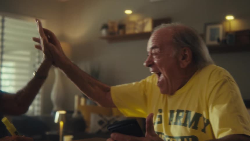 A man wearing a yellow us army tshirt and a watch sits at a table with a woman smiling with excitement as they high five and clasp hands