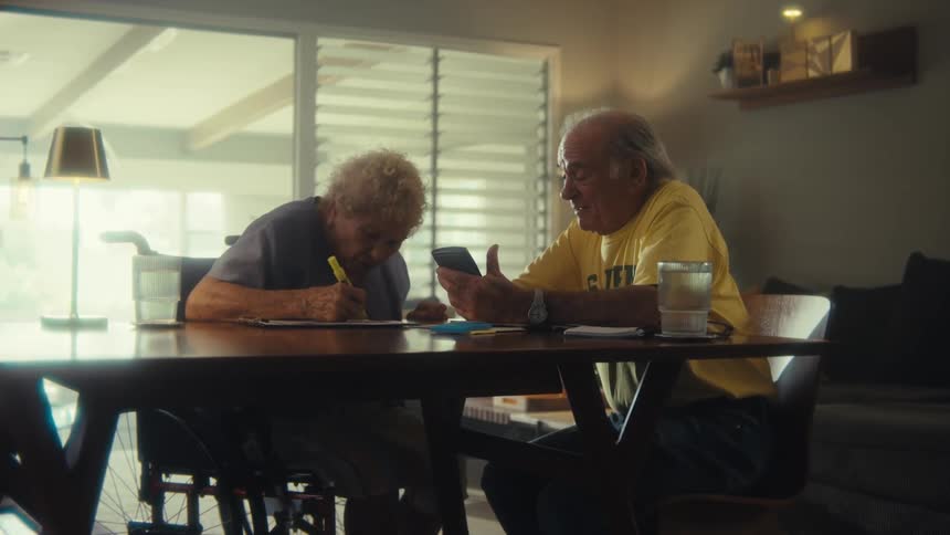 Two elderly individuals sitting at a dining table one engaged with a smartphone and the other writing in a sunlit home setting