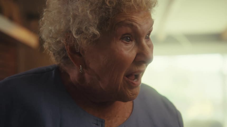 A closeup side profile of an elderly woman with white curly hair as her expression changes from surprise with an open mouth to a big joyful smile