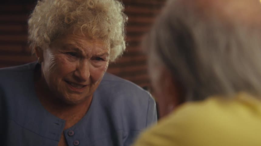 A close view of a woman with curly white hair wearing a light blue collared shirt as she talks to a person across from her with her expression changing from serious to a big smile