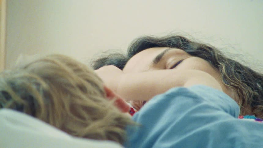 A close view of two people lying down and looking at each other with a woman on the right with dark hair smiling and a child with light colored hair on the left