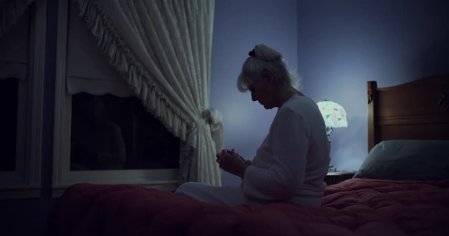 An old woman sitting on the edge of her bed at night praying while lit by a lamp on her nightstand