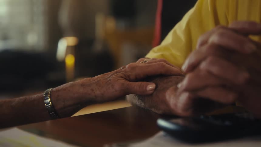 An elderly couple sits behind a desk in the living room holding hands at midday