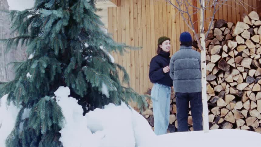 A teenage couple picks up firewood stacked beside a building the girl places the wood on the boys arm then kisses him as they walk through the snow at midday both wearing winter clothing