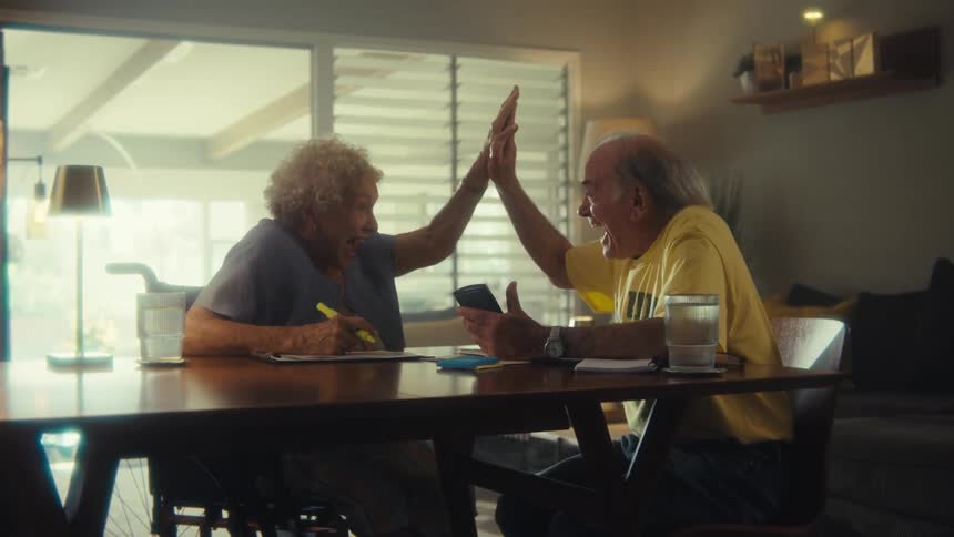 An elderly man and woman sitting at a table laughing and touching hands