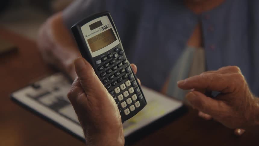A very close view shows a persons wrinkled hands holding a black calculator and pressing a button holding an elderly woman hand