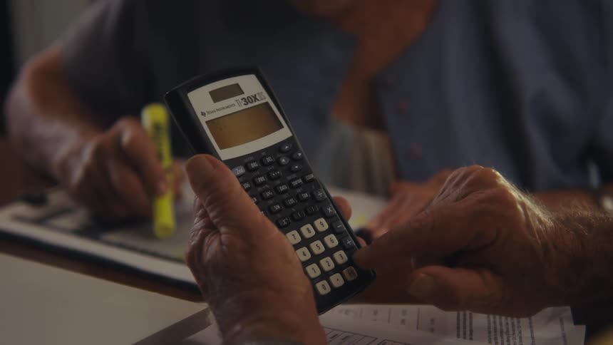 An elderly person holds a calculator while another woman beside him uses a marker to write on a document indoors
