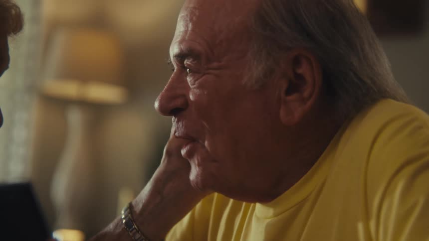 A man with graying hair and a yellow shirt sits across a table from a woman with curly white hair as they talk and he smiles